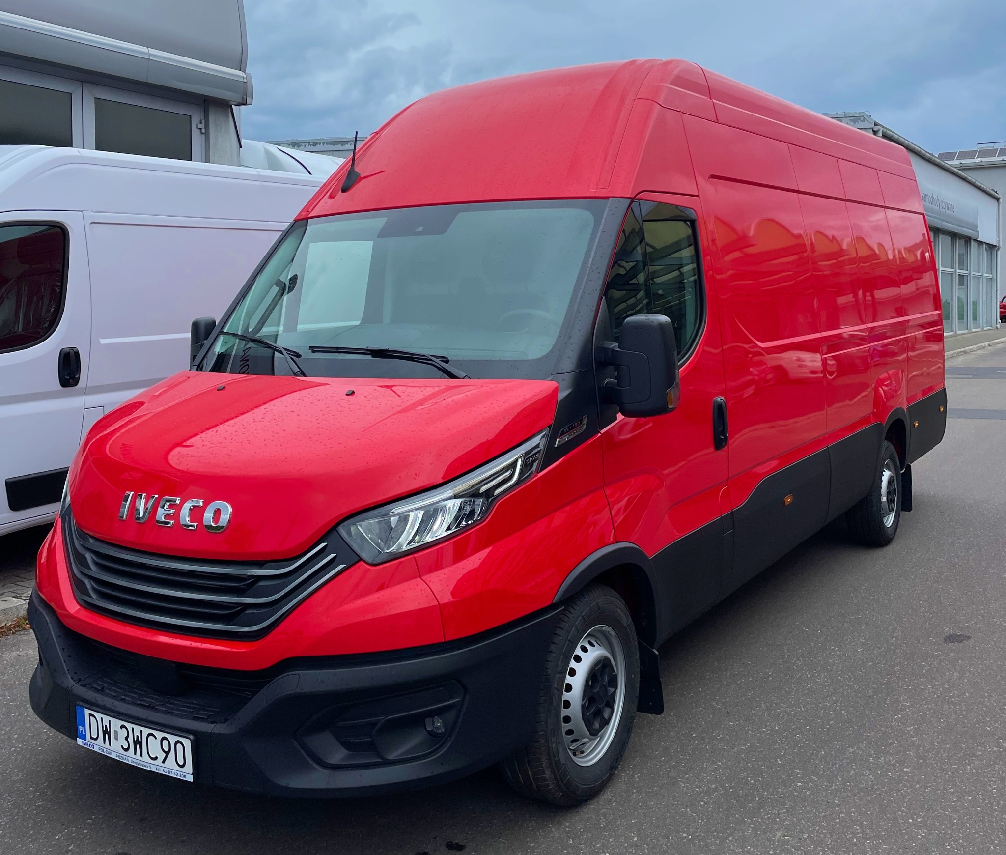 Czerwony, fabrycznie nowy furgon Iveco Daily z wysokim dachem, zaparkowany na zewnątrz salonu samochodowego w pochmurny dzień.