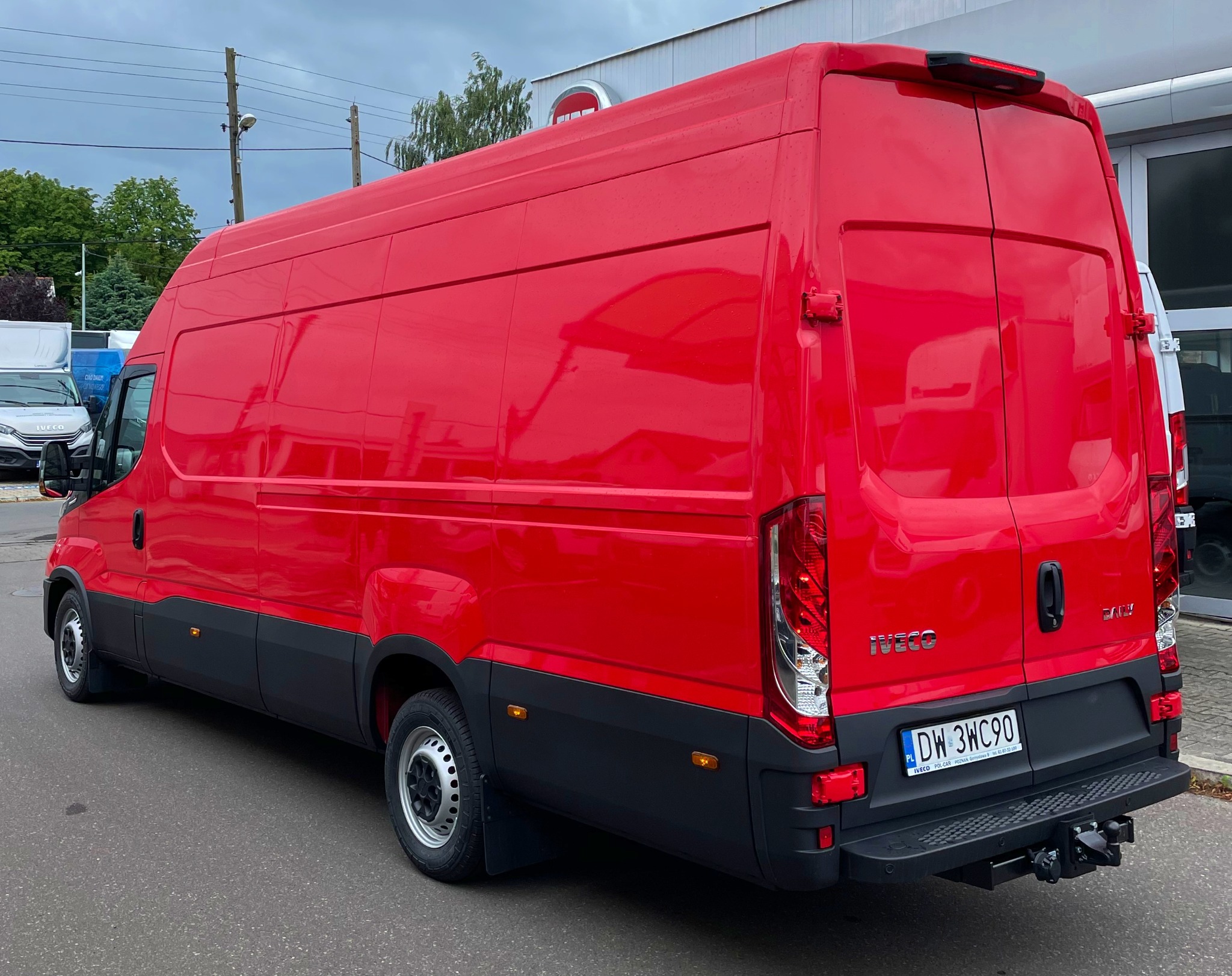 Czerwony samochód dostawczy Iveco Daily z polską tablicą rejestracyjną DW 3WC90, widok z tyłu pod kątem, na parkingu przed budynkiem firmy.