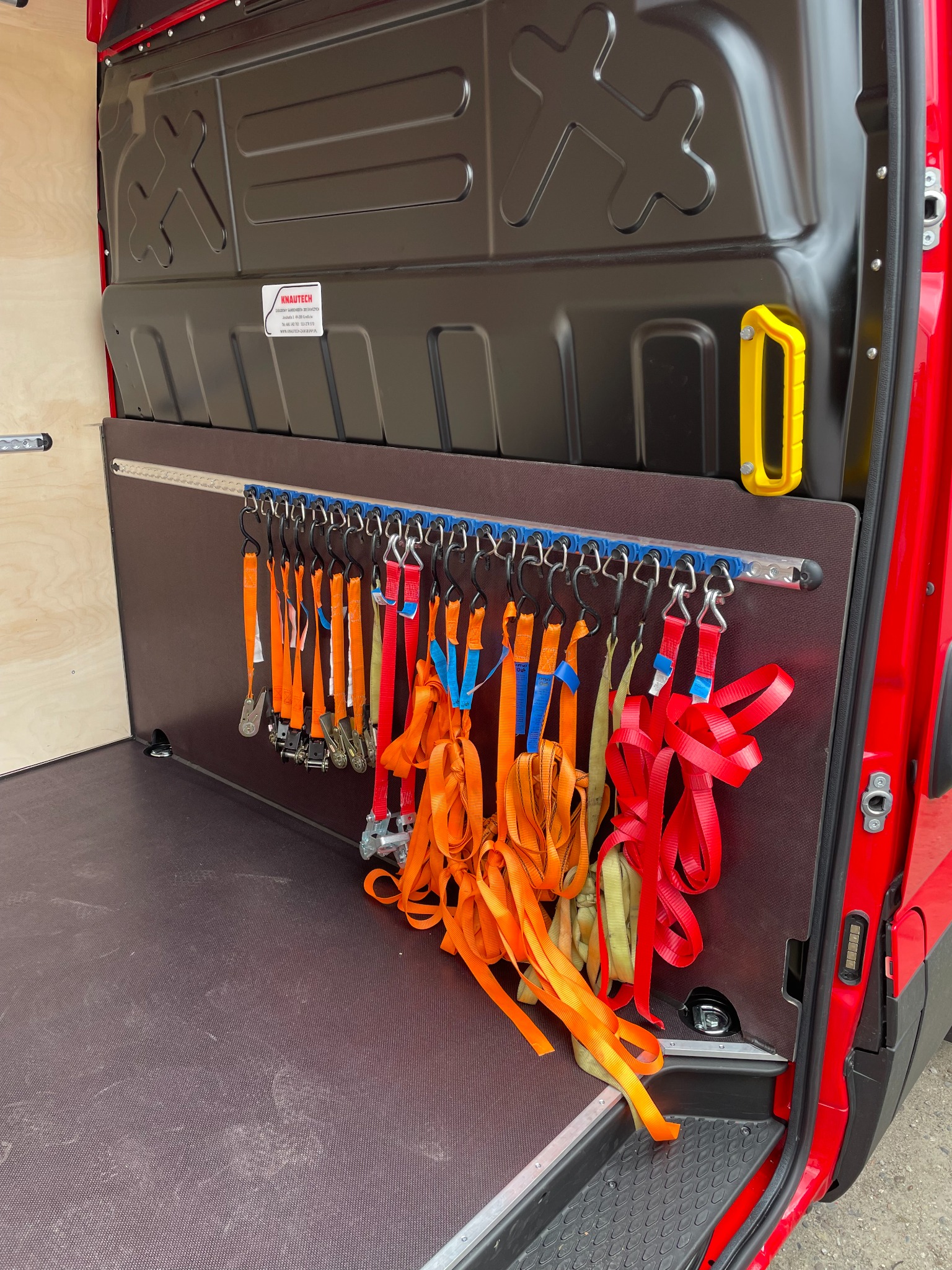 Wnętrze czerwonego busa z zamontowanymi zabezpieczeniami ładunku: szyny mocujące z haczykami i różnokolorowe pasy transportowe zwisające z nich.