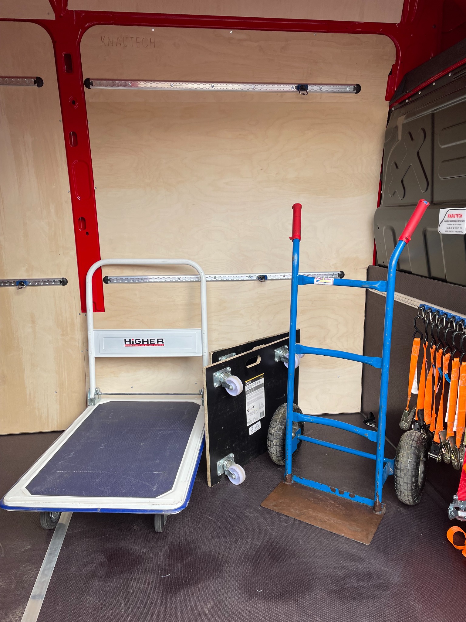 Wnętrze furgonetki z zamontowanymi szynami mocującymi, wózek platformowy marki Higher i wózek transportowy z czerwonymi uchwytami oraz pasy transportowe na wieszakach.