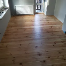 Wycyklinowana podłoga z jasnego drewna w pokoju z szarymi ścianami, widoczny grzejnik i okno, ślady pyłu na parapecie, niebieska taśma malarska przy futrynie.