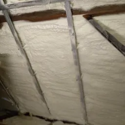 Poddasze z widocznymi krokwiami i metalowymi profilami, pokryte grubą warstwą białej pianki izolacyjnej o nieregularnej strukturze, z fragmentem płyty OSB w lewym dolnym rogu.