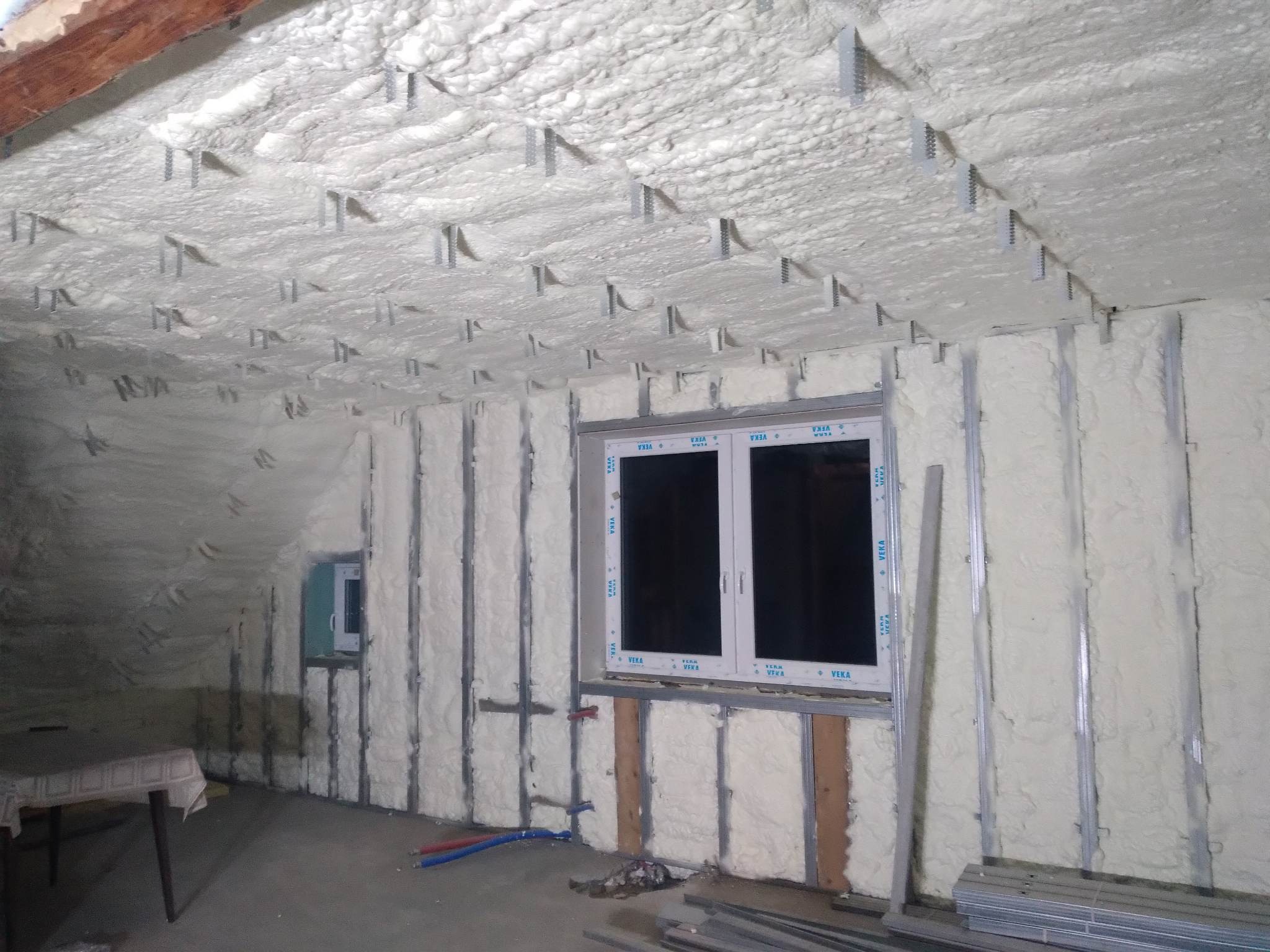 Poddasze w trakcie ocieplania pianką PUR, widoczne metalowe profile montażowe na ścianach i suficie, białe okno PCV z szybami zespolonymi, oraz fragment stołu z białym obrusem w rogu pomieszczenia.