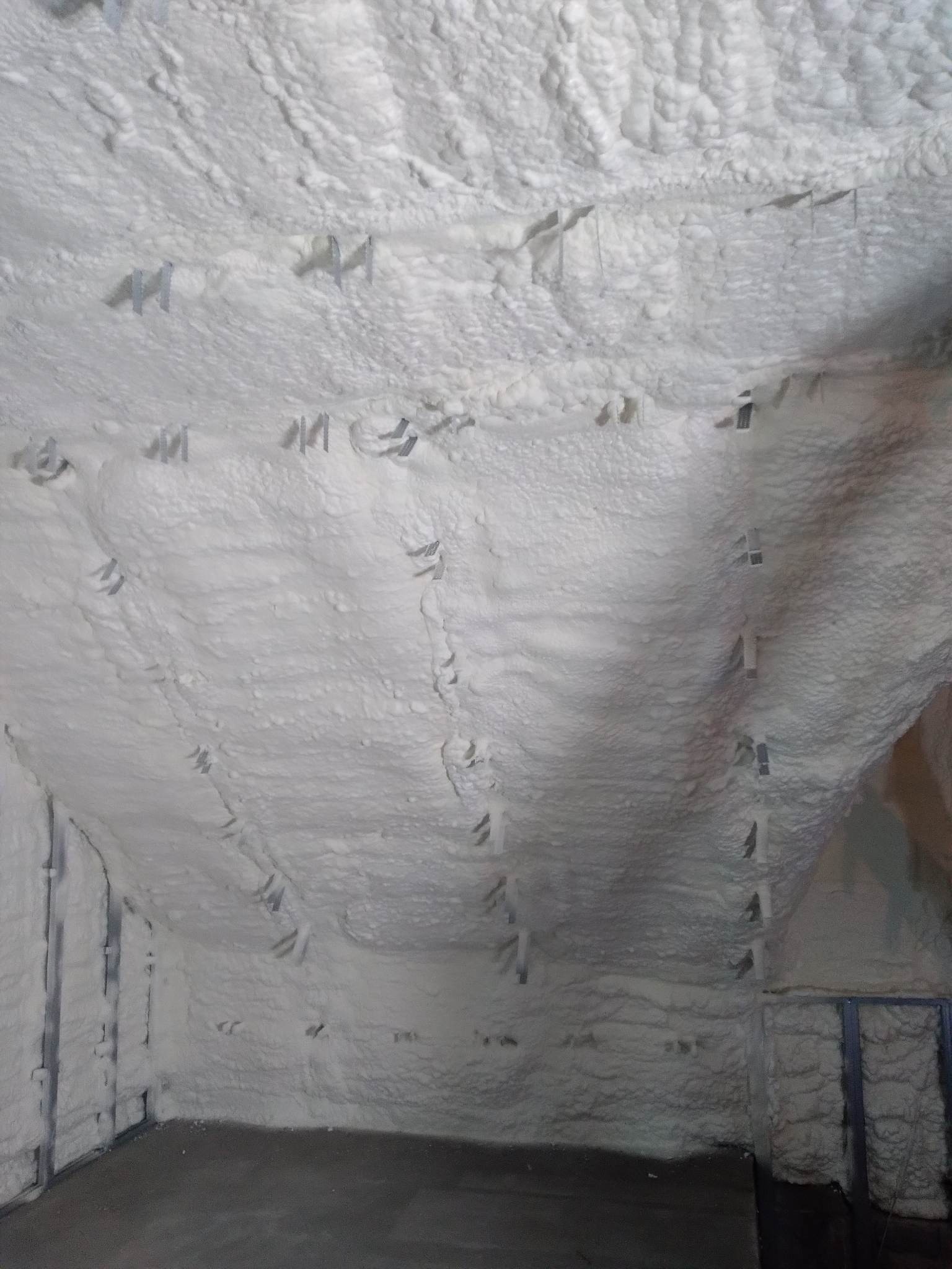 Poddasze po ociepleniu białą pianką poliuretanową, widoczne metalowe elementy mocujące w warstwie izolacyjnej, szara betonowa podłoga.