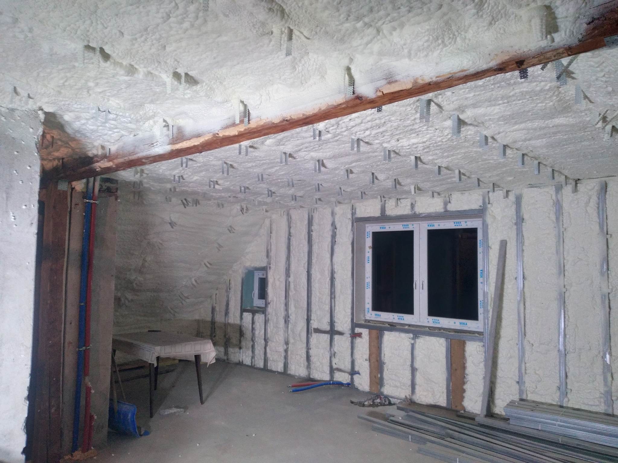 Poddasze po natrysku pianki poliuretanowej, widoczne elementy konstrukcyjne dachu i okna, fragment stołu z lewej strony, instalacja elektryczna na ścianie.
