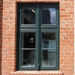 Dwuskrzydłowe okno z ciemnozielonymi ramami osadzone w elewacji z czerwonej cegły z dekoracyjnym łukowym nadprożem z cegieł ułożonych pionowo.