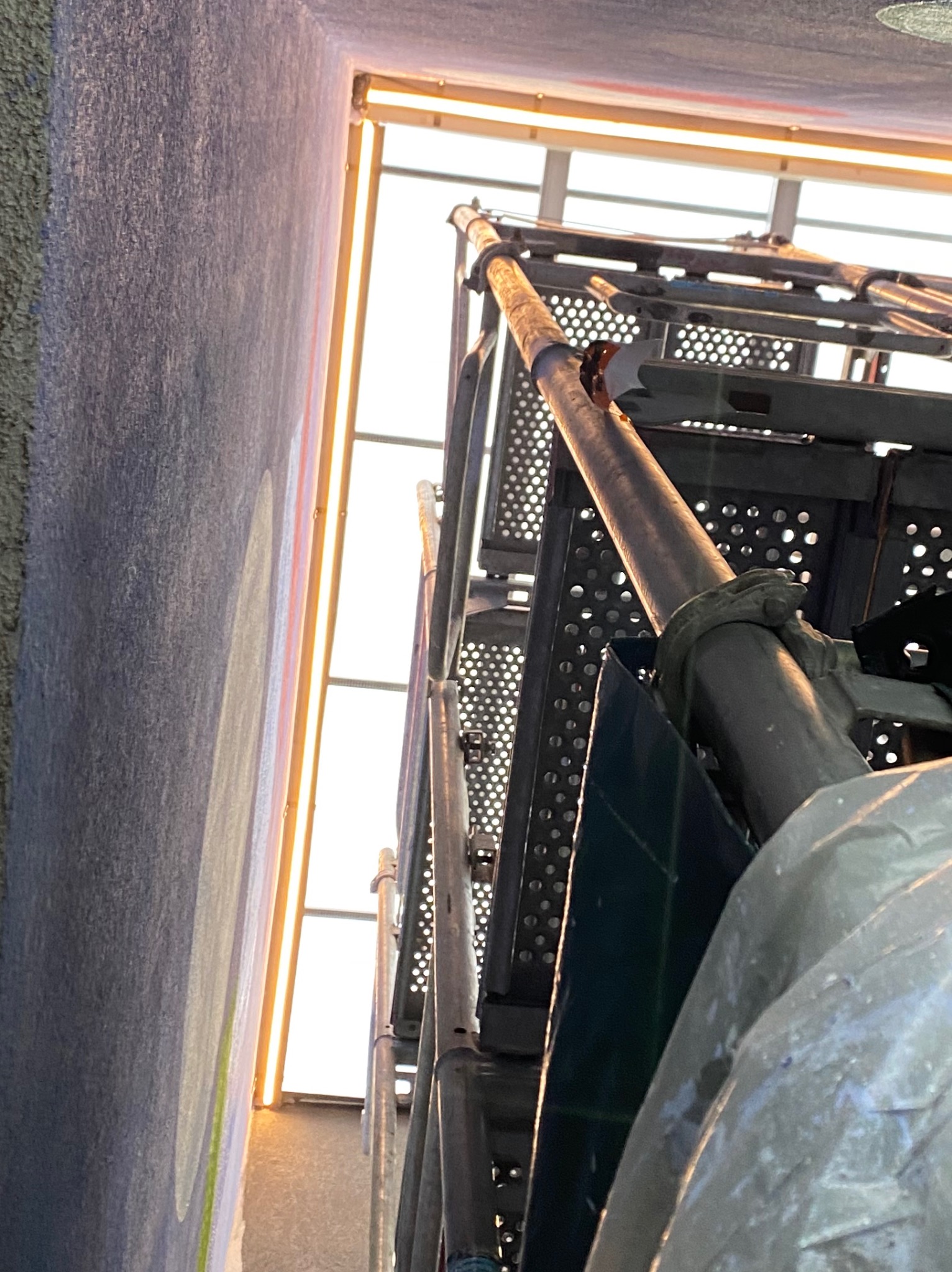 Wysoki kąt widzenia rusztowania ustawionego przy ścianie budynku z oknami, widoczne elementy mocujące i metalowa perforowana podłoga rusztowania.