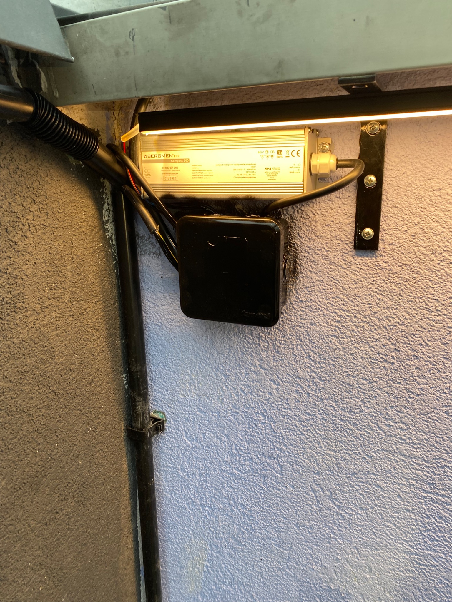 Zasilacz LED marki Bergmen Eco Compact 200, zamontowany na szorstkiej, niebieskiej ścianie, z widocznymi przewodami w peszlu i listwą LED. Widoczne śruby mocujące i tabliczka znamionowa urządzenia.