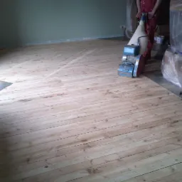 Cyklinowanie drewnianej podłogi maszyną z widocznymi śladami po szlifowaniu, fragmentem płytek podłogowych przy ścianie i meblami zabezpieczonymi folią.