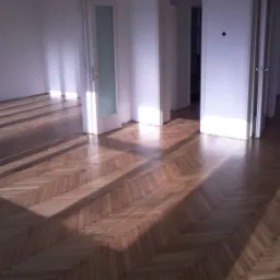 Parkiet ułożony w jodełkę po cyklinowaniu, z widocznymi promieniami słońca wpadającymi przez okno, podkreślającymi strukturę drewna i gładką powierzchnię.