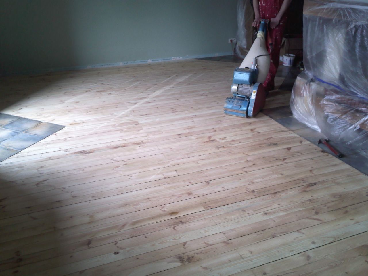 Cyklinowanie drewnianej podłogi maszyną z widocznymi śladami po szlifowaniu, fragmentem płytek podłogowych przy ścianie i meblami zabezpieczonymi folią.