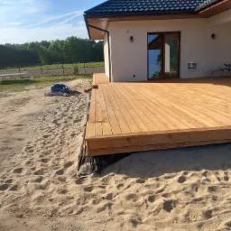 Jasny, drewniany taras przylegający do nowoczesnego domu z ciemnym dachem, otoczony piaszczystym terenem i zielenią w tle. Na piasku widoczny zwinięty niebieski wąż ogrodowy.