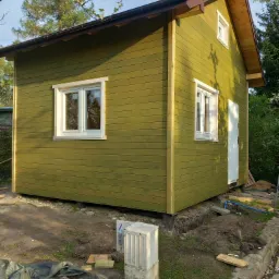 Nowoczesny, zielony domek letniskowy z drewnianą elewacją, białymi oknami i brązowym dachem, widziany z boku w słoneczny dzień, z widocznymi elementami placu budowy.