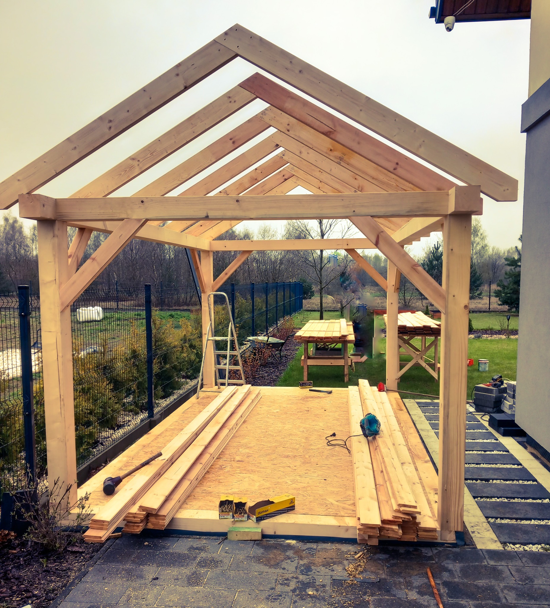 Konstrukcja drewniana altany w trakcie budowy, widoczna więźba dachowa, słupy nośne, podłoże z płyt OSB, przygotowane deski, narzędzia i drabina w tle.