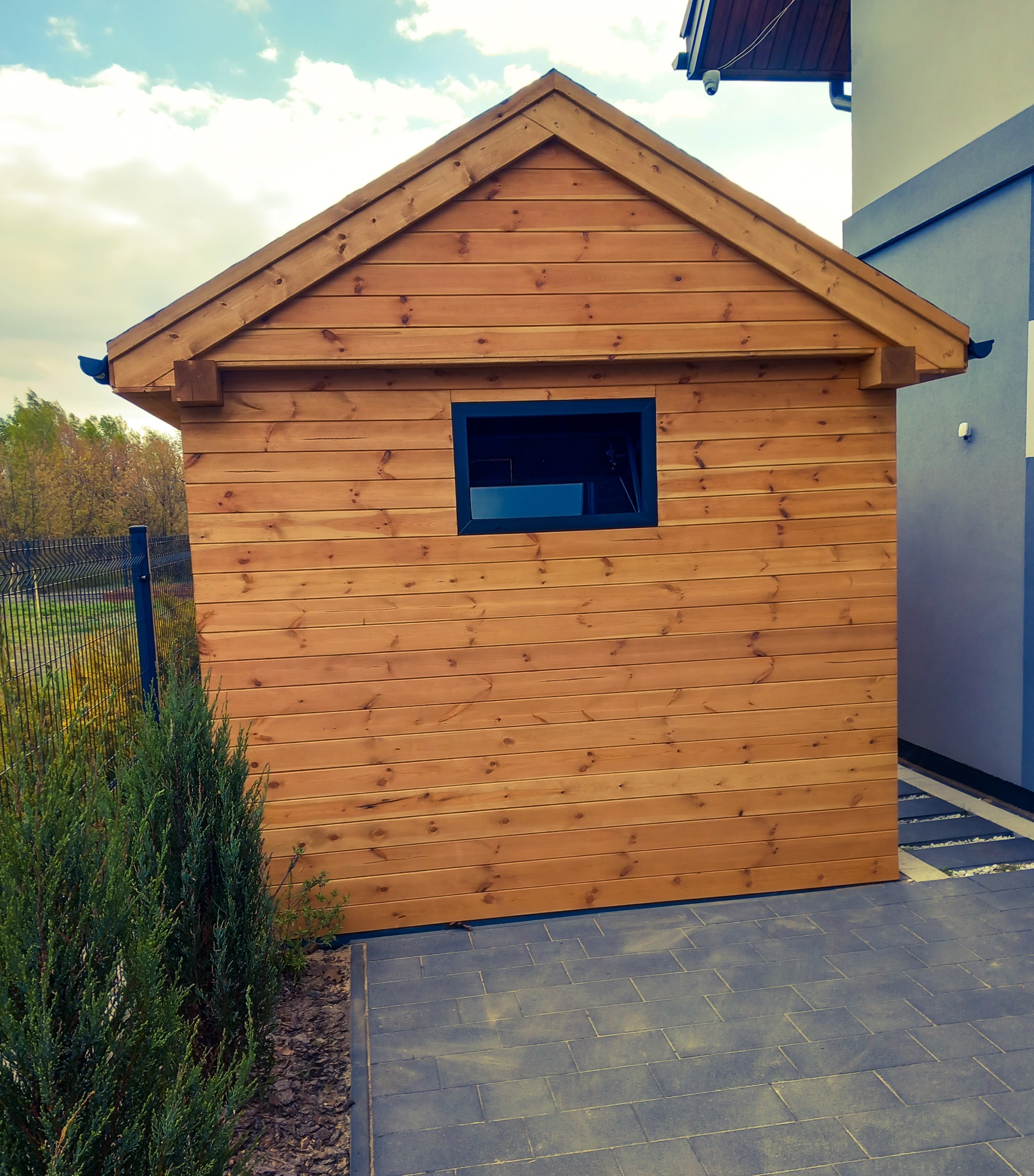 Drewniana konstrukcja z oknem, prawdopodobnie domek narzędziowy, z widoczną fakturą drewna i czarną ramą okienną, na tle kostki brukowej i zieleni.