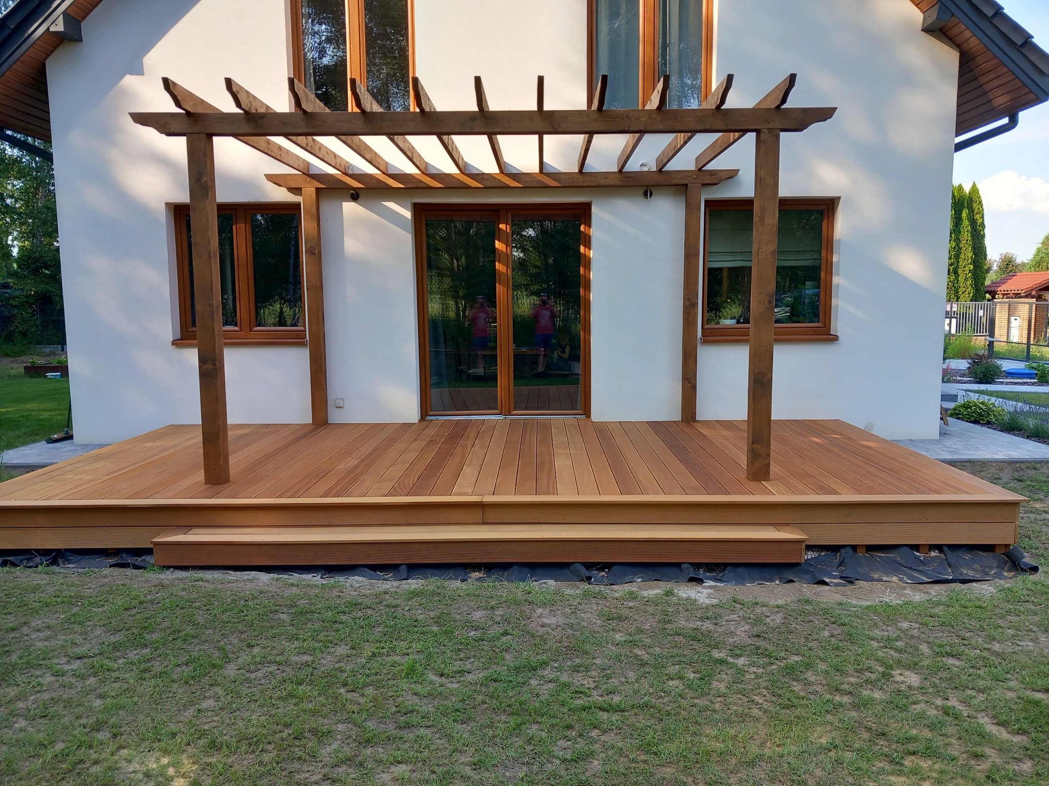 Nowoczesny taras drewniany z pergolą w odcieniach brązu, przylegający do białego domu z dużymi oknami, widoczny czarny materiał pod konstrukcją tarasu.