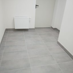 A.S.Bud Oleksandr Sokhmii - Szare, matowe płytki podłogowe ułożone w korytarzu, widoczny biały grzejnik i listwy przypodłogowe w kolorze płytek.