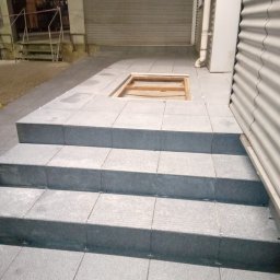 A.S.Bud Oleksandr Sokhmii - Schody i fragment posadzki wyłożone szarymi płytkami, widoczne krzyżyki dystansowe, w tle brama garażowa i fragment budynku z blachy falistej, otwór w posadzce bez wykończenia.