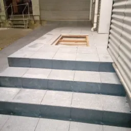 Schody i fragment posadzki wyłożone szarymi płytkami, widoczne krzyżyki dystansowe, w tle brama garażowa i fragment budynku z blachy falistej, otwór w posadzce bez wykończenia.