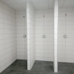 A.S.Bud Oleksandr Sokhmii - Trzy kabiny prysznicowe wyłożone białymi, prostokątnymi płytkami, z szarą podłogą i widocznymi przyłączami wody (czerwone i niebieskie) oraz zamontowanymi bateriami.