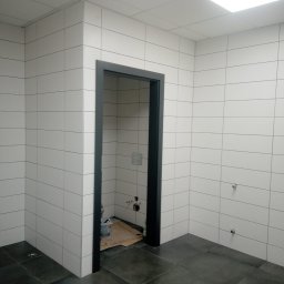 A.S.Bud Oleksandr Sokhmii - Wnętrze łazienki w trakcie prac wykończeniowych: białe, prostokątne płytki ścienne z ciemnoszarą fugą, ciemnoszara podłoga z płytek, czarna ościeżnica drzwiowa, widoczne przyłącza wodne na ścianie,...