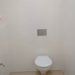 A.S.Bud Oleksandr Sokhmii - Nowoczesna, minimalistyczna toaleta z białymi, poziomymi płytkami i drewnopodobną podłogą, widoczny przycisk spłuczki w kolorze szarym.