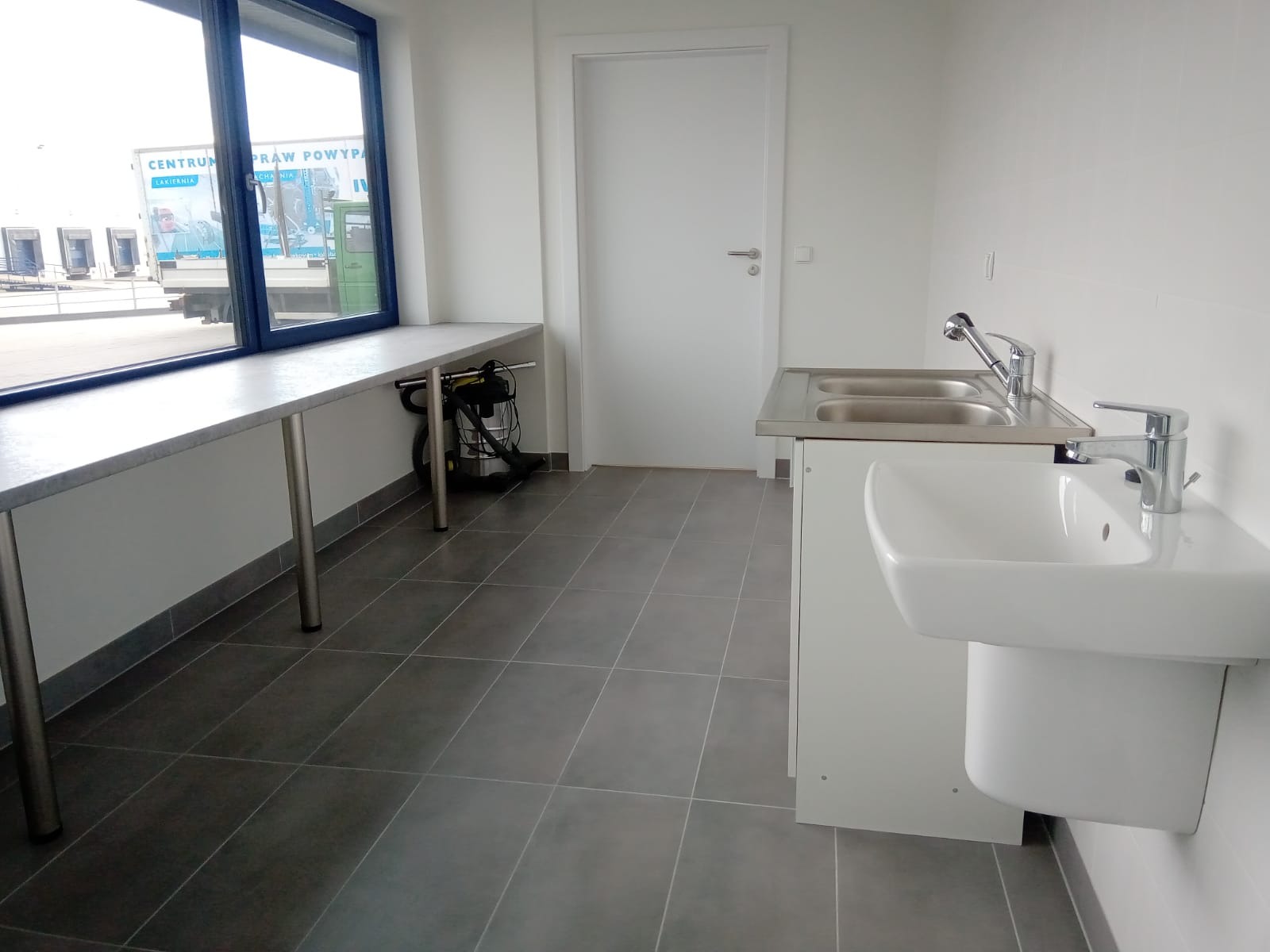 Nowoczesna, minimalistyczna łazienka w biurze z szarymi płytkami podłogowymi, białą umywalką z podwójnym zlewem i chromowanymi bateriami, z oknem wychodzącym na plac załadunkowy i odkurzaczem...