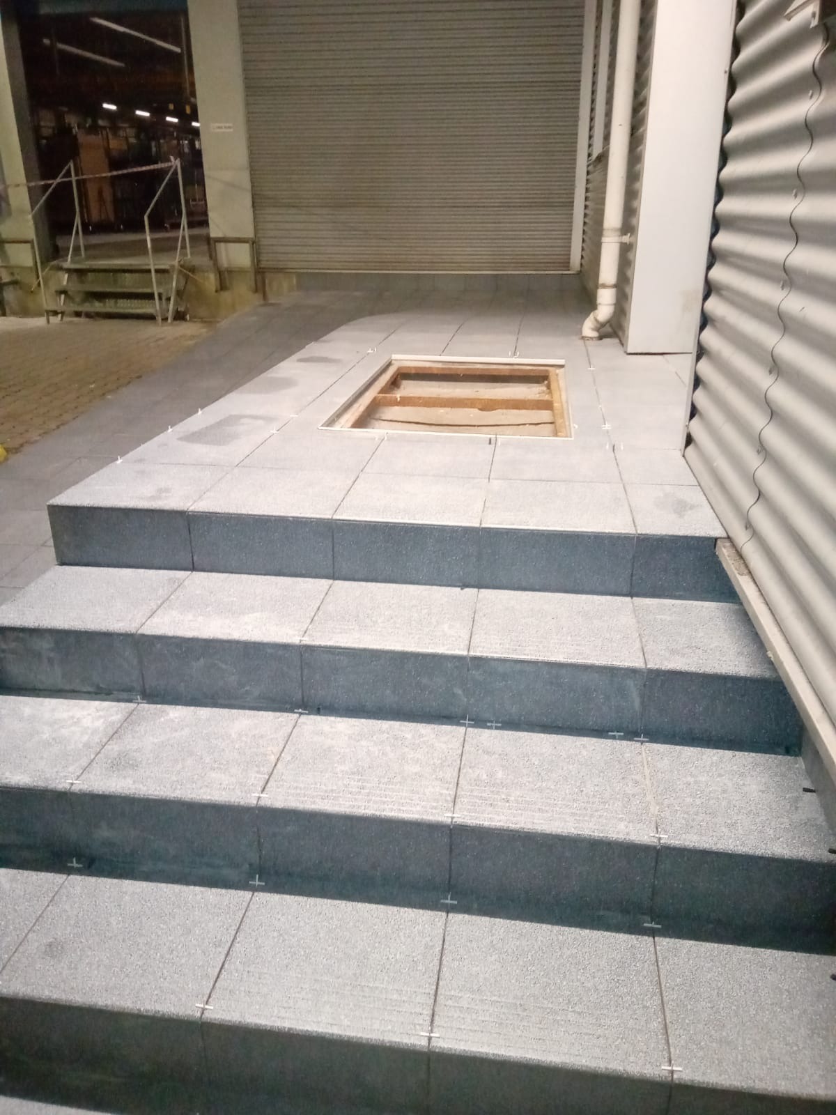 Schody i fragment posadzki wyłożone szarymi płytkami, widoczne krzyżyki dystansowe, w tle brama garażowa i fragment budynku z blachy falistej, otwór w posadzce bez wykończenia.