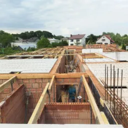 Wznoszenie ścian z czerwonej cegły na budowie domu, widok z góry na strop Teriva, drewniane szalunki i zbrojenia, w tle zabudowa jednorodzinna i pochmurne niebo.