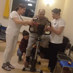 Starszy mężczyzna na rowerze stacjonarnym, wspierany przez dwie kobiety i obserwowany przez dziecko w domowym otoczeniu.