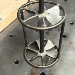 Metalowa konstrukcja z centralnym prętem i wirnikiem wewnątrz okrągłej ramy, spawana na perforowanym metalowym stole warsztatowym, z fragmentami maszyny w tle.