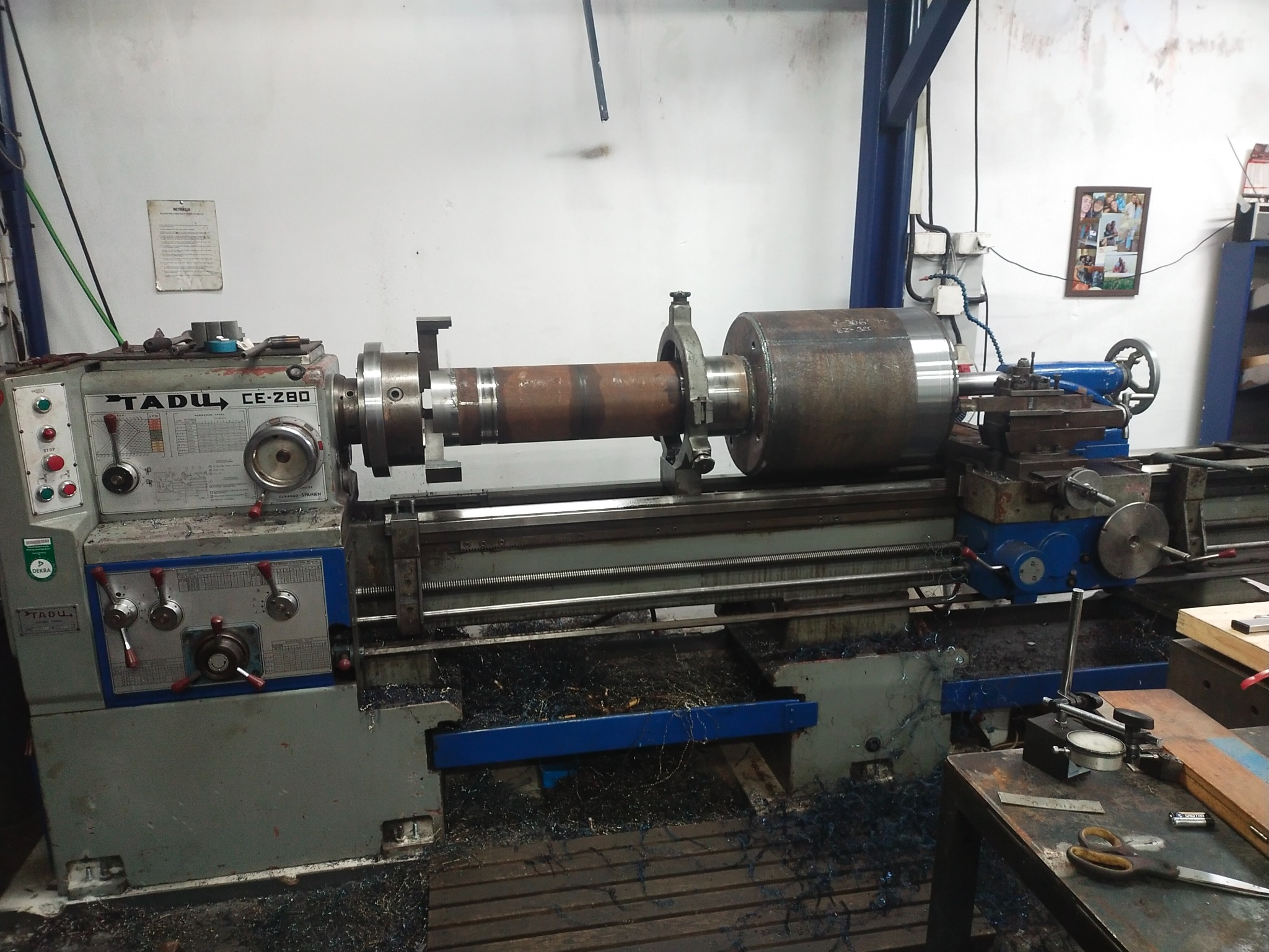 Ciężka tokarka Tadu CE-280 w warsztacie, obrabiająca duży, rdzawy, cylindryczny element z metalu, wióry na podłożu, narzędzia pomiarowe na stole obok.