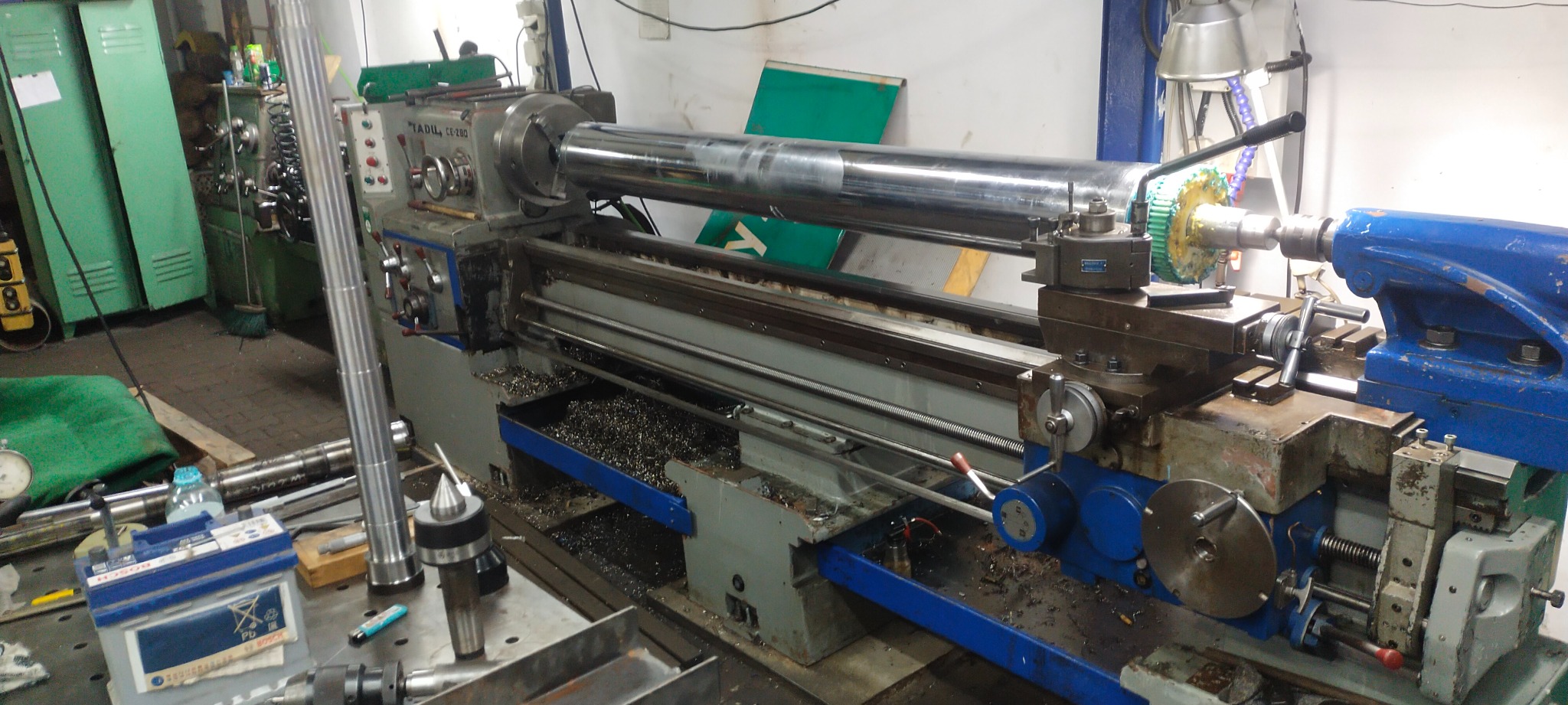 Długi, chromowany wał obrabiany na tokarce TADIL CE-280, wióry metalowe widoczne w korycie, narzędzia i akcesoria na stole roboczym.