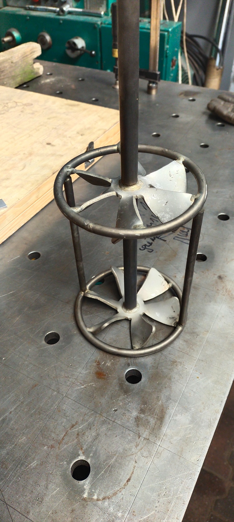Metalowa konstrukcja z centralnym prętem i wirnikiem wewnątrz okrągłej ramy, spawana na perforowanym metalowym stole warsztatowym, z fragmentami maszyny w tle.