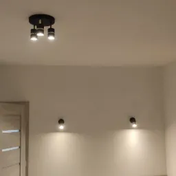 Nowoczesne oświetlenie sufitowe i ścienne w nowym domu w Ostrowie Wielkopolskim, widoczna ściana z odsłoniętą cegłą obok okna.