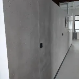Długa, szara ściana z efektem betonu architektonicznego, w tle widoczne biuro z przeszkleniami i czarnymi włącznikami światła.