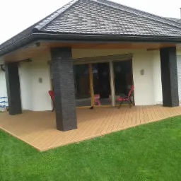 Zadaszony taras z desek kompozytowych, podtrzymywany przez ceglane kolumny, z widokiem na zadbany trawnik i dom z szarym dachem.