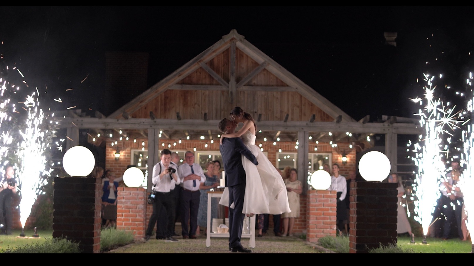 Nowożeńcy w objęciach na tle drewnianej konstrukcji, otoczeni gośćmi i fontannami iskier podczas wieczornego przyjęcia weselnego.