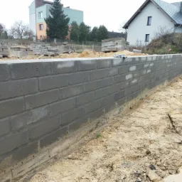 Wznoszenie ściany fundamentowej z szarych bloczków betonowych na piaszczystym podłożu, widoczne palety z bloczkami w tle.