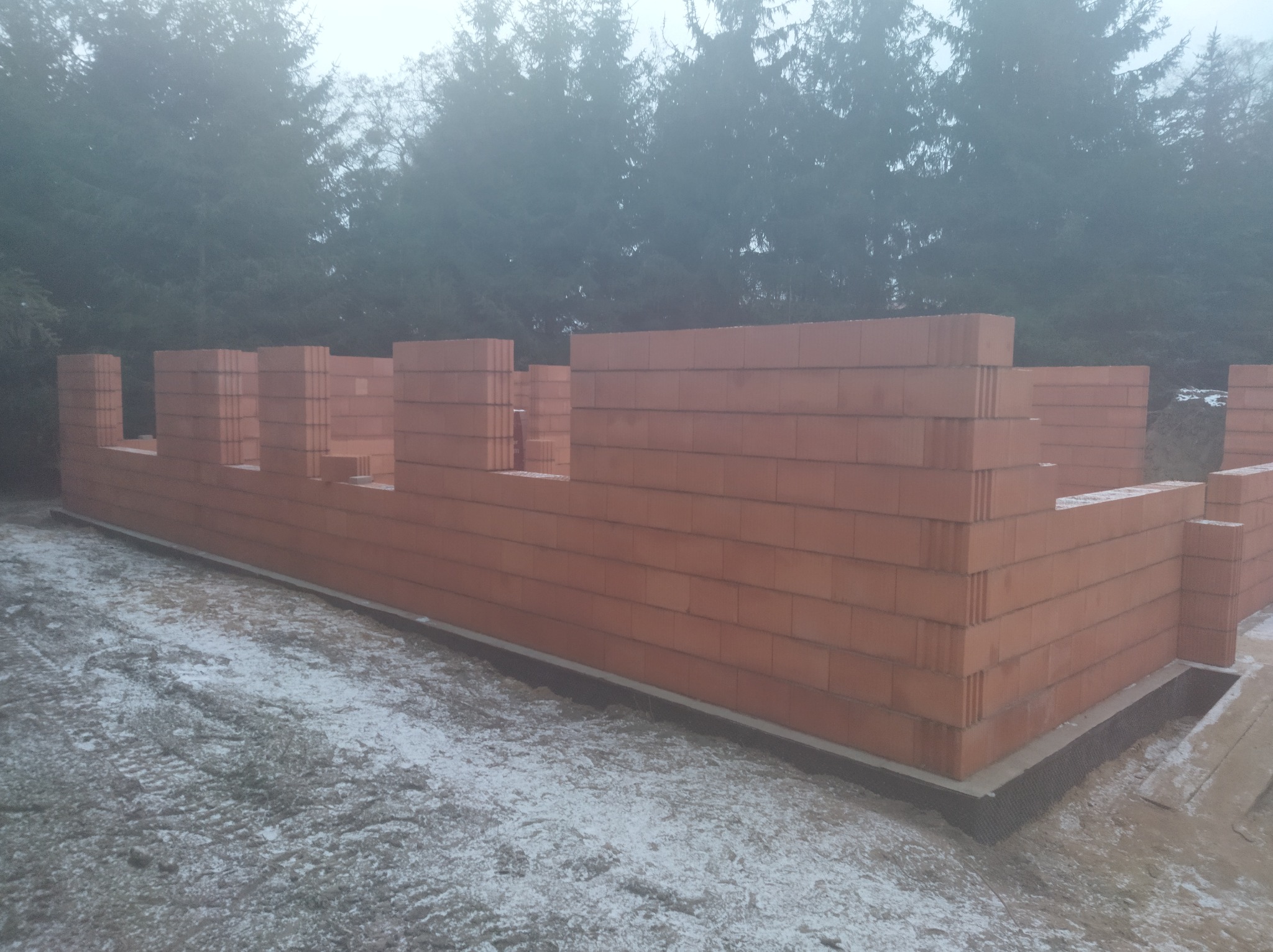 Wznoszone ściany z czerwonej cegły ceramicznej na fundamencie, widoczne fragmenty ośnieżonej ziemi i zarys drzew we mgle w tle.