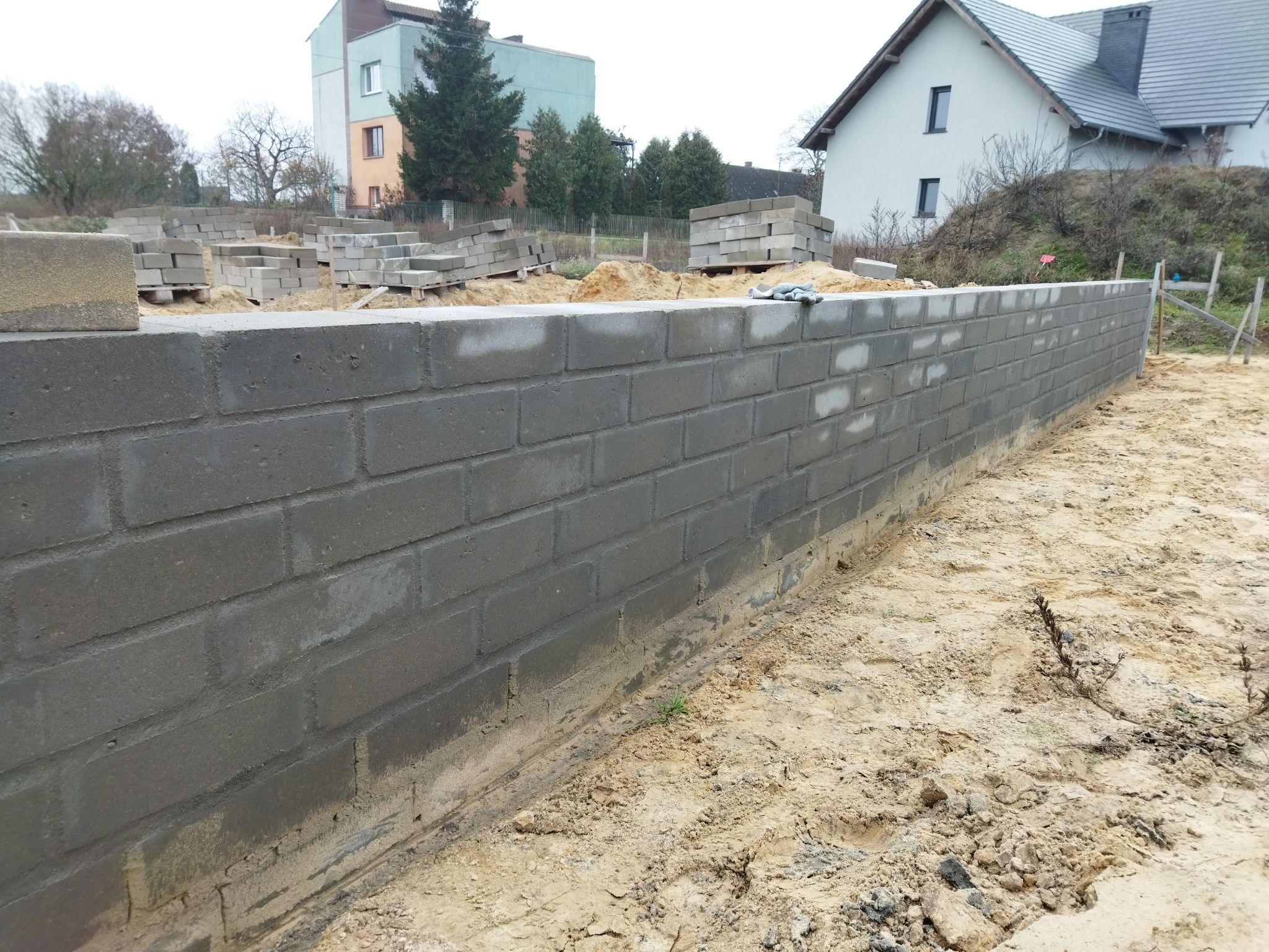 Wznoszenie ściany fundamentowej z szarych bloczków betonowych na piaszczystym podłożu, widoczne palety z bloczkami w tle.