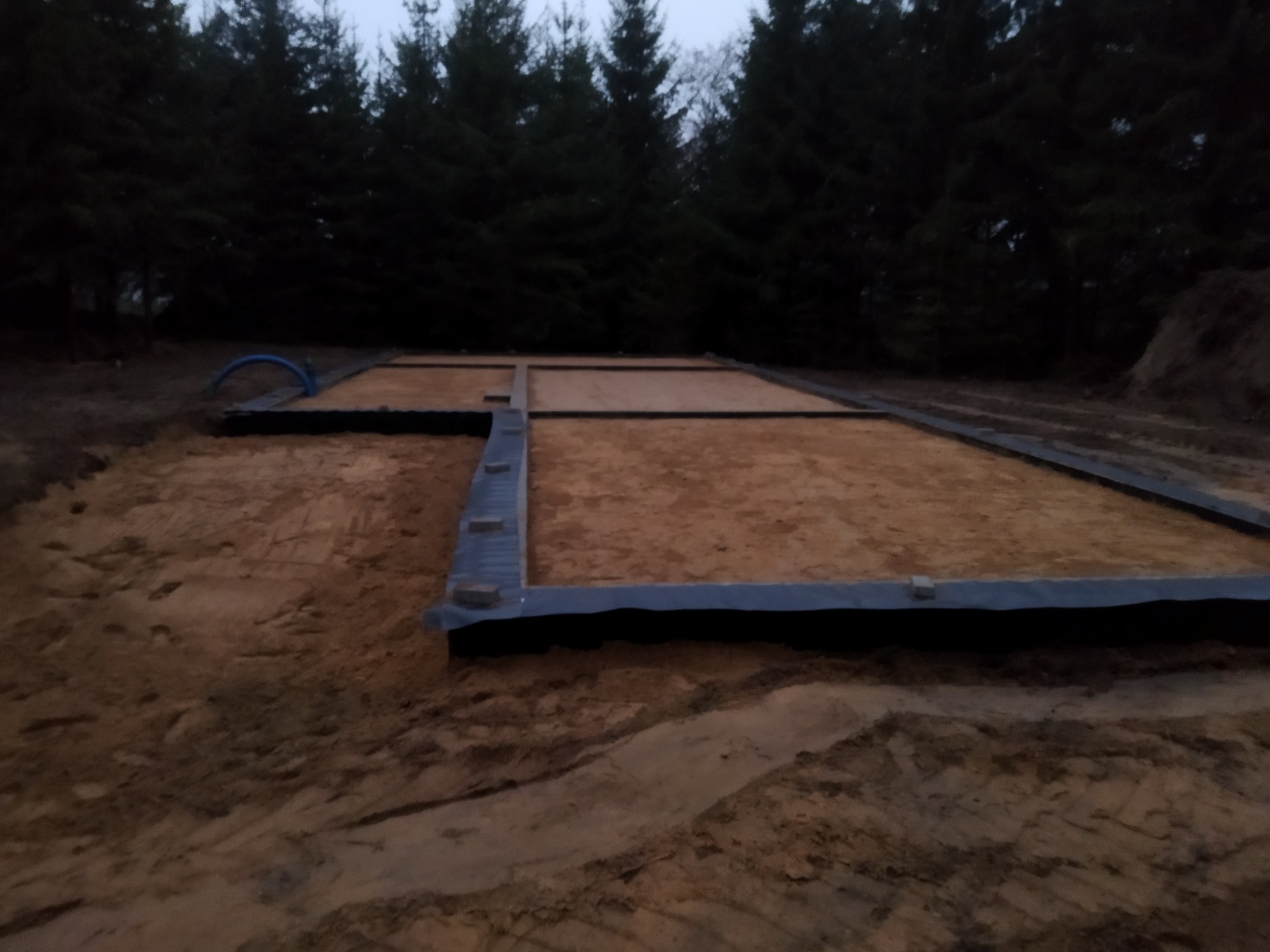 Przygotowany fundament pod budowę domu z izolacją w ziemi, widok na teren budowy z drzewami w tle.