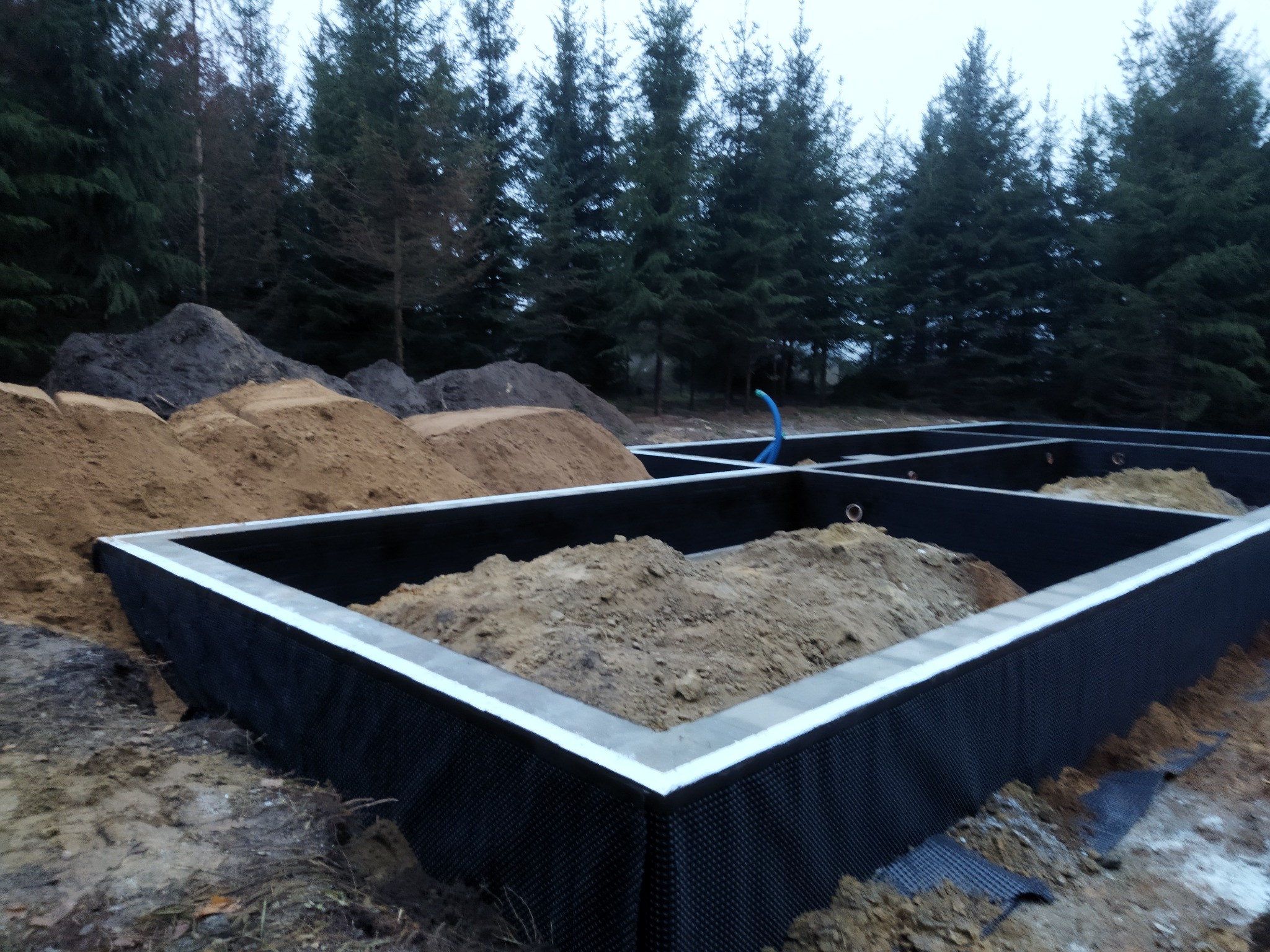 Fundamenty domu murowanego wypełnione ziemią, widoczne zabezpieczenie przeciwwilgociowe, w tle las iglasty i hałdy ziemi.