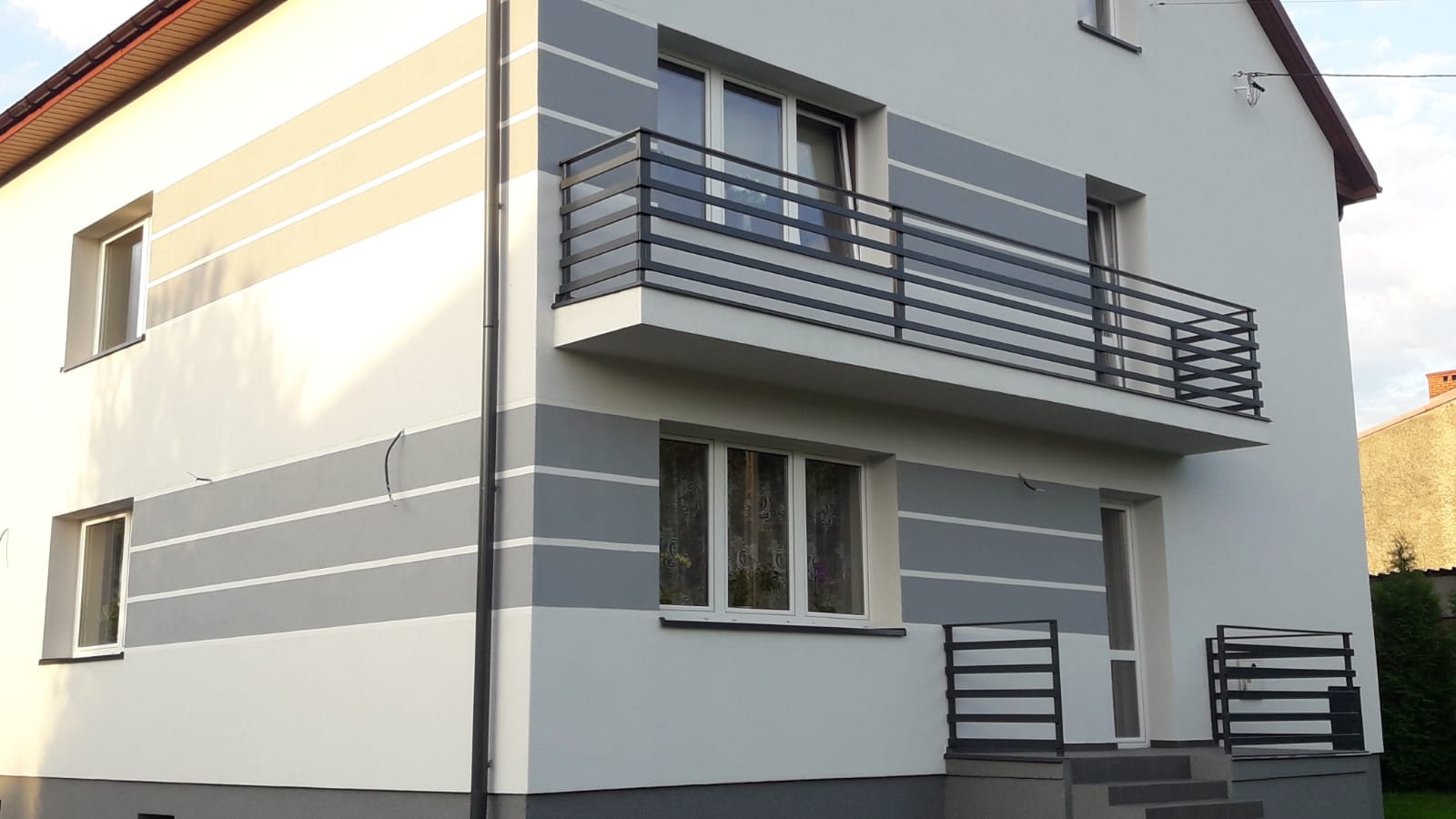 Nowoczesny dom z balkonem zabezpieczonym czarną, poziomą balustradą, dopasowaną do poręczy schodów wejściowych i szarego pasa na elewacji.