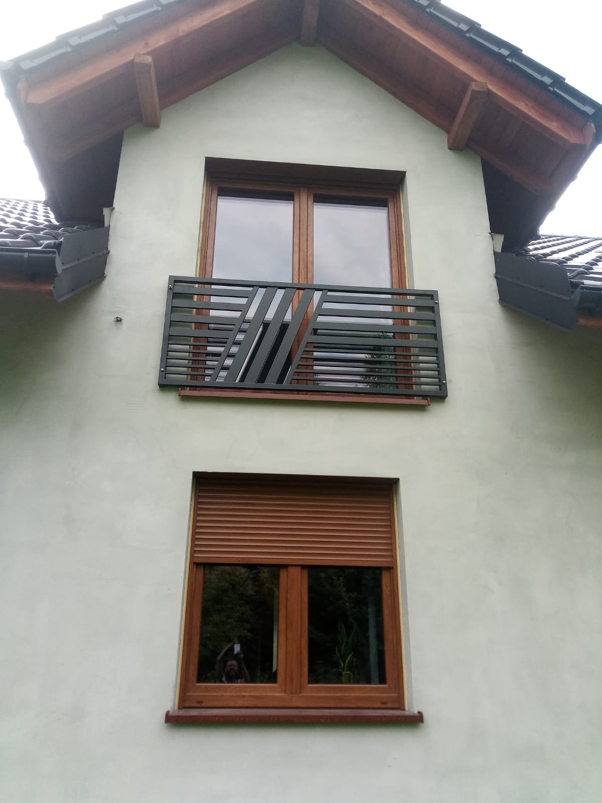 Nowoczesna, grafitowa barierka balkonowa z diagonalnym wzorem na tle jasnej elewacji domu z drewnianymi oknami i roletami zewnętrznymi.