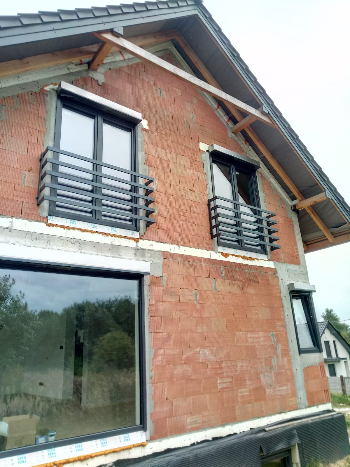 Surowy budynek w trakcie budowy z zamontowanymi czarnymi balustradami okiennymi o prostej konstrukcji, widoczne okna z roletami zewnętrznymi.