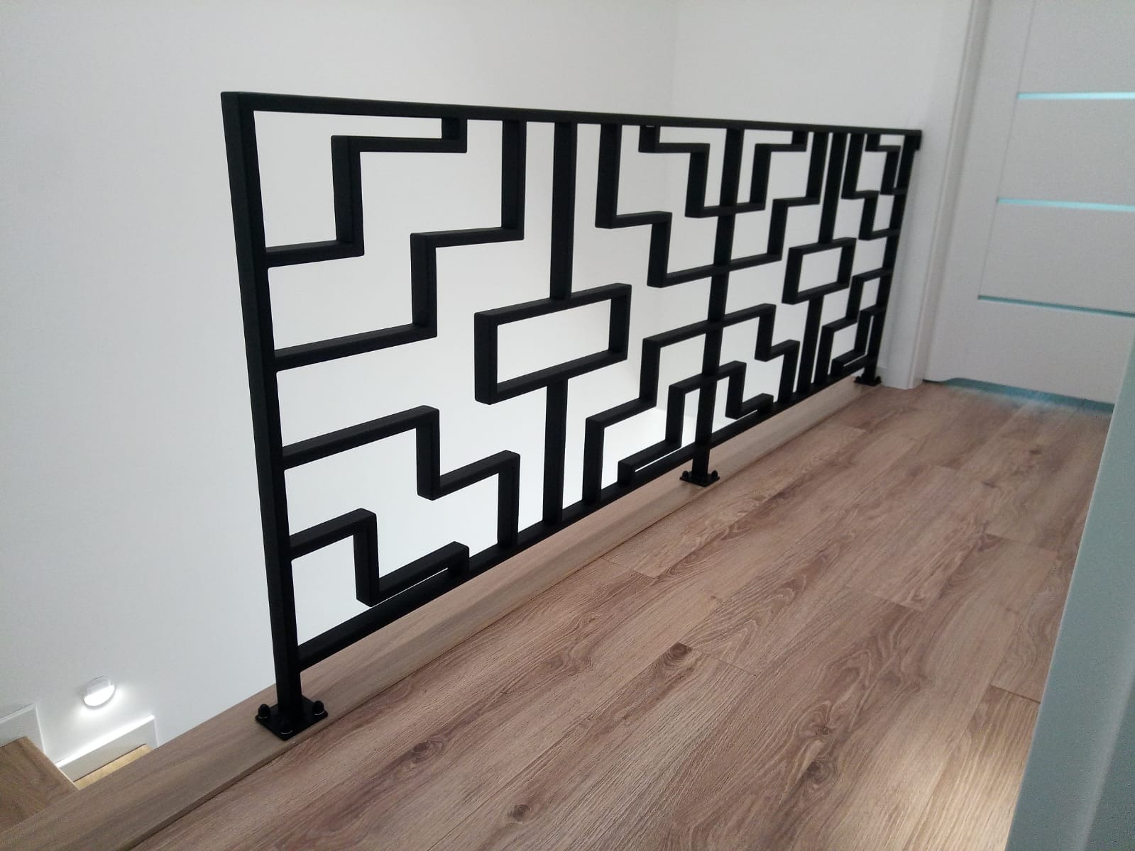 Czarna, metalowa barierka z geometrycznym wzorem na drewnianej podłodze, nowoczesny design.