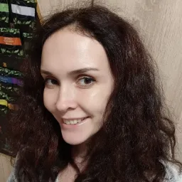 Uśmiechnięta kobieta z brązowymi, kręconymi włosami, pozująca na tle szafki z herbatami.