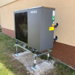 Szara pompa ciepła Heiko Thermal Plus zamontowana na metalowej konstrukcji na betonowych podstawach, z widocznymi rurami wchodzącymi do budynku o dwukolorowej elewacji.
