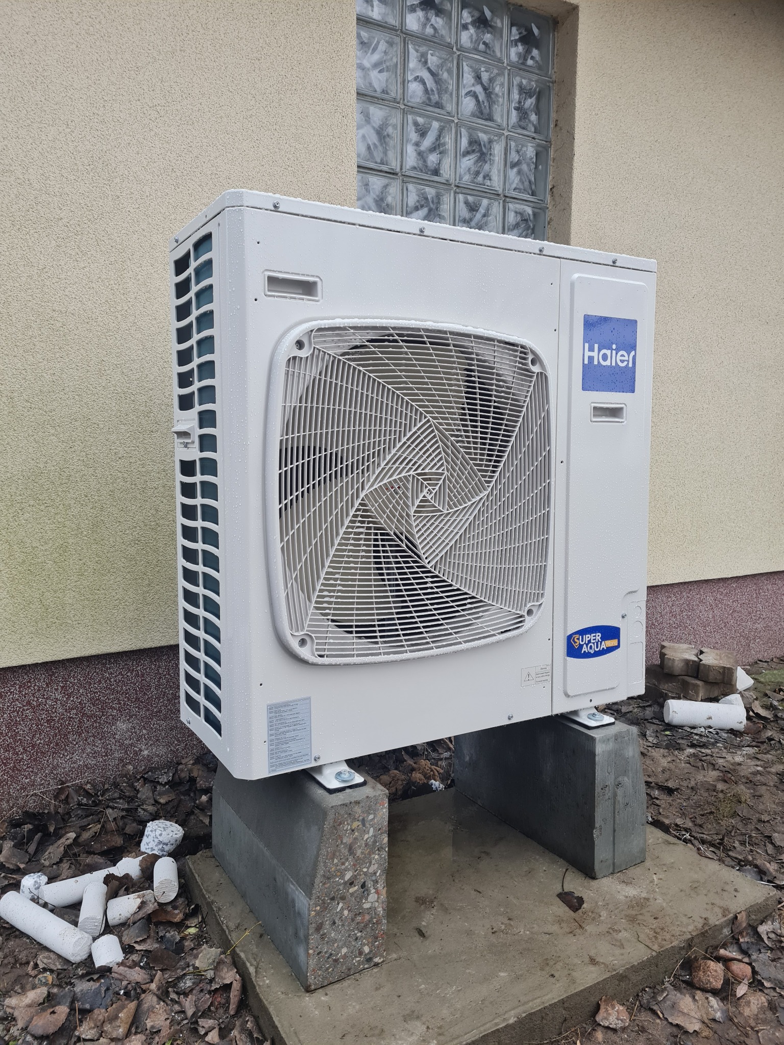 Biała zewnętrzna jednostka pompy ciepła Haier Super Aqua, umieszczona na betonowych blokach przy ścianie budynku z oknem z luksferów, widoczne ślady wilgoci i resztki materiałów izolacyjnych.